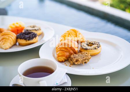 Un cadre de petit déjeuner avec une tasse de thé, des croissants, des beignets et un muffin sur des assiettes blanches près d'une piscine. Banque D'Images