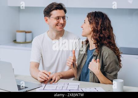 Couple souriant assis à table dans leur cuisine, examinant les plans et la disposition de la nouvelle maison. L'homme, portant des lunettes et une chemise blanche, tient un stylo, tandis que la femme, avec les cheveux bouclés et la veste verte, donne les pouces vers le haut. Banque D'Images