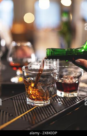 Gros plan d'une main versant du whisky ambré d'une bouteille dans un verre de roches dans un bar avec des lumières bokeh Banque D'Images