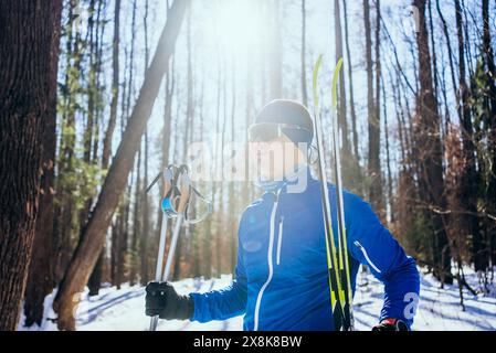 L'homme se tient confiant dans la forêt ensoleillée, prêt pour le ski de fond. Il porte une veste bleue, des lunettes de soleil et un bandeau, tenant des bâtons de ski et des skis. Banque D'Images