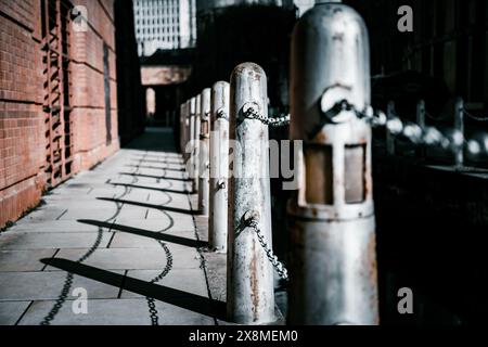 Un sentier urbain avec des poteaux et des chaînes métalliques, de longues ombres. Les bâtiments en briques créent une atmosphère industrielle et gritty. Banque D'Images