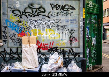 Manchester UK 26 mars 2024. Mur de la ville recouvert de graffitis colorés, entouré de sacs poubelles débordants et de boîtes en carton dans une ruelle urbaine. Banque D'Images