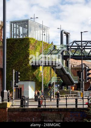 Manchester UK 26 mai 2024. Cadre urbain moderne avec jardin vertical, pont piétonnier, ascenseur en verre, et les gens marchant sous un ciel bleu clair Banque D'Images