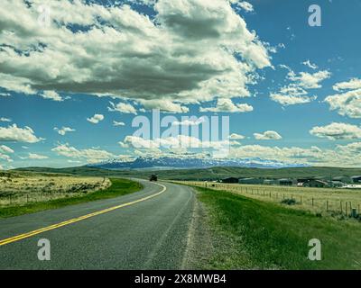 L'image montre les nuages et la route de l'autoroute courbant alors qu'elle se déplace vers la chaîne de montagnes Absaroka près de Cody, Wyoming. Le paysage est rural avec des bâtiments commerciaux. Banque D'Images