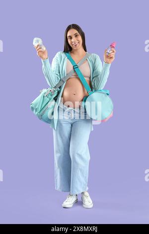 Jeune femme enceinte avec des sacs d'hôpital de maternité et des biberons sur fond lilas Banque D'Images