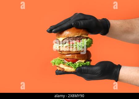 Mains masculines dans des gants noirs tenant de savoureux hamburgers sur fond orange Banque D'Images