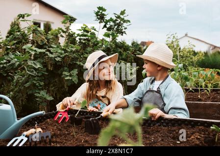 Fille et garçon prenant soin de petites plantes de légumes dans un lit surélevé, tenant une petite pelle. Enfance en plein air dans le jardin. Banque D'Images