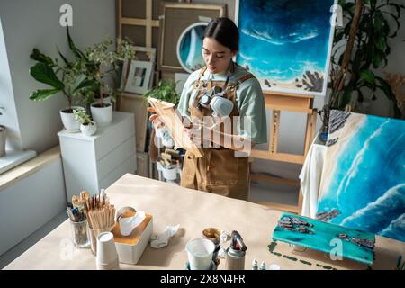 Artiste peintre féminin focalisé prépare un panneau de bois pour l'application de résine époxy dans un atelier d'art. Banque D'Images