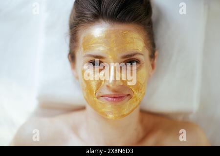 Temps de soins. Vue supérieure de la femme moderne détendue dans le salon de spa avec masque cosmétique doré sur le visage couché sur la table de massage. Banque D'Images