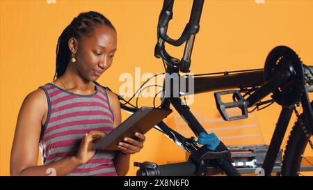 Femme afro-américaine faisant le service de vélo dans un atelier de réparation de fond de studio, regardant sur la liste d'entretien de tablette. Technicien vérifiant les composants de bicyclette qui doivent être réparés, caméra A. Banque D'Images