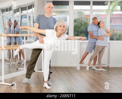 Femme âgée souriante portant des vêtements de sport dansant en couple et la formation ballet arabesque pose avec partenaire pendant le cours de groupe au salon de danse Banque D'Images