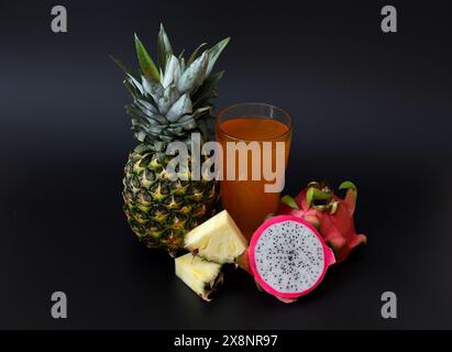 Un grand verre de jus fraîchement pressé sur un fond noir, à côté de morceaux d'ananas mûrs et de pitaya. Gros plan. Banque D'Images