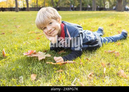 mignon garçon de 7 ans est allongé sur l'herbe par une chaude journée ensoleillée d'automne. joie, atmosphère positive, enfance heureuse. Bonjour, automne. Promenez-vous dans le parc Banque D'Images