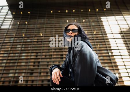 Jeune femme portant des lunettes de soleil et un sweat à capuche noir est accroupie, regardant avec confiance la caméra avec un bâtiment urbain moderne dans le dos Banque D'Images