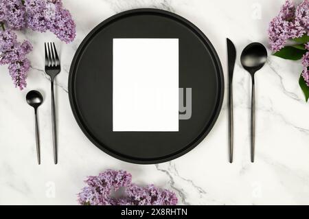 Vue de dessus de fleurs de lilas violettes sur fond de marbre blanc. Table de table, assiette noire et couverts, carte blanche. Papier peint de printemps, pose à plat, copie spac Banque D'Images