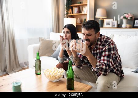 Jeune couple excité et tendu assis sur un canapé à la maison regardant ou diffusant des événements sportifs à la télévision ensemble. Homme et femme regardent le football ou le match de football Banque D'Images