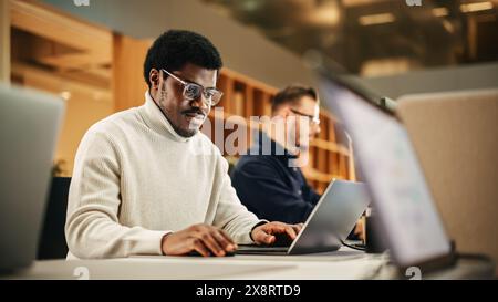 Portrait d'un homme noir enthousiaste tournant sur son ordinateur portable et commençant sa journée de travail au bureau. Graphiste masculin souriant tout en développant un nouveau concept visuel Banque D'Images