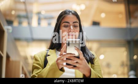 Portrait d'une jeune femme hispanique utilisant son smartphone, marchant dans un hall de bureau occupé et souriant. Prise de vue moyenne. Une femme d'affaires en vêtements décontractés intelligents entre dans son lieu de travail le matin Banque D'Images