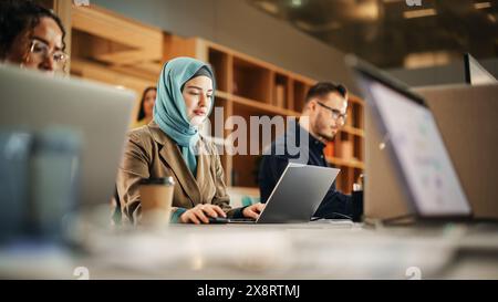 Portrait de femme musulmane focalisée utilisant un ordinateur portable dans un bureau spacieux contemporain. Spécialiste des comptes souriant tout en ayant une journée productive. Collègues actifs travaillant sur des ordinateurs en arrière-plan. Banque D'Images