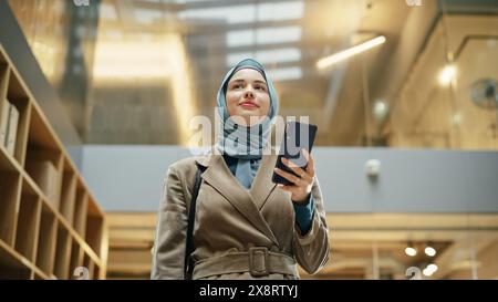 Portrait de femme d'affaires portant Hijab en utilisant l'application smartphone pour partager social Media Post sur la croissance de carrière. Gestionnaire professionnelle femme marchant à travers l'immeuble de bureaux, souriant et regardant heureux. Banque D'Images