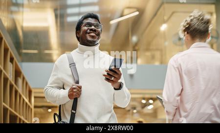 Portrait du jeune homme noir se promène dans le couloir du siège social, en utilisant son smartphone et en souriant. Homme d'affaires prospère communiquant avec ses collègues par SMS. Prise de vue à angle bas Banque D'Images