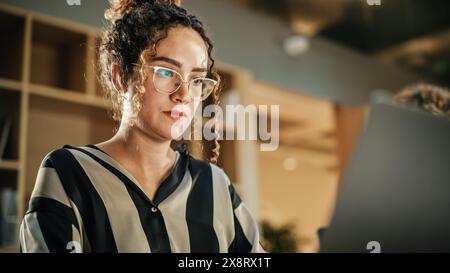 Portrait d'hispanique inspiré jeune femme souriant et utilisant un ordinateur portable dans un bureau contemporain lumineux. Conseillère cliente répondant aux e-mails du service clientèle, yeux concentrés sur l'écran. Gros plan. Banque D'Images