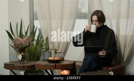 Homme caucasien détendu dans l'intérieur de la cuisine moderne appréciant le thé et utilisant la tablette. Support. Jeune freelance ayant une pause du travail à la maison. Banque D'Images