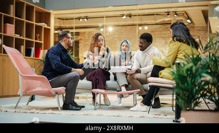 Groupe multiethnique de personnes créatives travaillant sur projet dans un bureau lumineux moderne. Un groupe diversifié de collègues développant une nouvelle approche à l'aide de graphiques, d'ordinateurs portables et de tablettes. Grand angle Banque D'Images