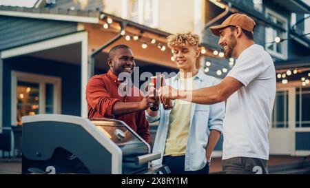 Jeune homme dans une casquette montrant son barbecue moderne à deux compagnons divers lors d'une Garden barbecue Party. Amis parlant, se relaxant, buvant des bières dans une arrière-cour Home Gathering. Banque D'Images