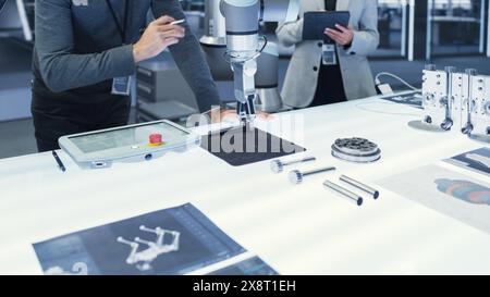 Bras robotique moderne tenant et déplaçant un composant mécanique métallique. Équipe diversifiée d'ingénieurs utilisant un ordinateur tablette pour manipuler et programmer le robot. Banque D'Images