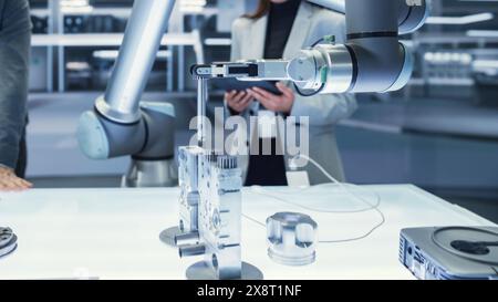 Gros plan d'un bras robotique moderne tenant et déplaçant un composant mécanique métallique. Équipe diversifiée d'ingénieurs utilisant un ordinateur tablette pour manipuler et programmer le robot. Banque D'Images