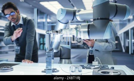 Gros plan d'un bras robotique moderne tenant et déplaçant un composant mécanique métallique. Équipe diversifiée d'ingénieurs utilisant un ordinateur tablette pour manipuler et programmer le robot. Banque D'Images