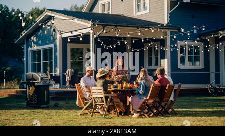 Grande famille et amis célébrant dehors à la maison. Groupe diversifié d'enfants, d'adultes et de personnes âgées réunis à une table, ayant des conversations amusantes. Préparer le barbecue et manger des légumes. Banque D'Images