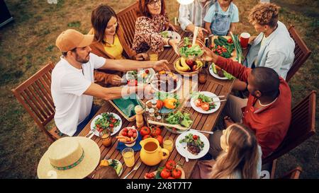 Vue surélevée de haut en bas d'une famille et des amis célébrant à l'extérieur à la maison. Groupe diversifié d'enfants, d'adultes et de personnes âgées assis à une table, ayant des conversations amusantes. Manger barbecue et légumes. Banque D'Images