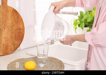 Femme versant de l'eau de la cruche filtrante dans le verre dans la cuisine, gros plan Banque D'Images