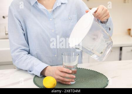 Femme versant de l'eau de la cruche filtrante dans le verre à la table en marbre blanc dans la cuisine, gros plan Banque D'Images