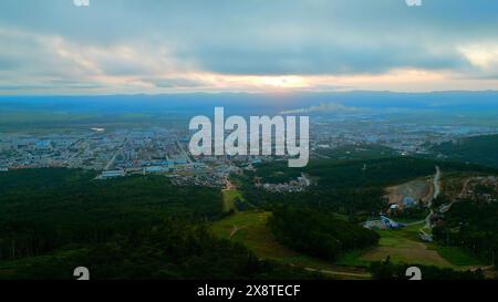 Vol aérien descendant les montagnes verdoyantes vers la ville touristique. Agrafe. Arbres verts d'été sur une pente de colline et la ville avec ciel nuageux au-dessus. Banque D'Images