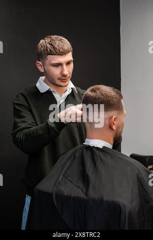 Jeune coiffeur coupe soigneusement les cheveux de Mans avec une tondeuse dans le salon de coiffure animé Banque D'Images