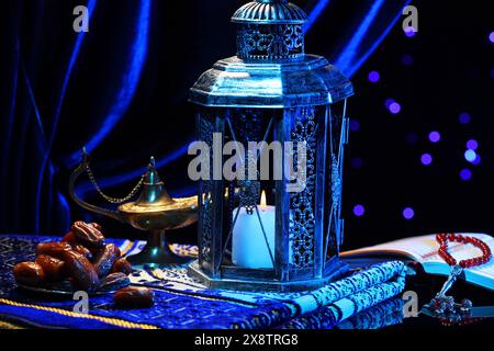 Lanterne arabe, Coran, misbaha, lampe magique Aladdin, dattes et tapis de prière plié sur la surface miroir contre les lumières floues la nuit Banque D'Images