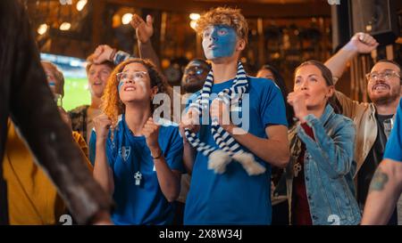 Groupe diversifié de fans de football aux visages colorés regardant un match de football en direct dans un bar sportif. Les gens encouragent leur équipe. Le joueur marque un but et la foule célèbre gagner le Championnat. Banque D'Images