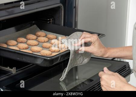mains de la personne utilisant un gant de four pour retirer un plateau de biscuits d'un four Banque D'Images