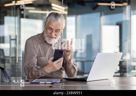 Dans un bureau moderne, un homme d'affaires âgé utilise un smartphone et un ordinateur portable, analysant des données, effectuant un appel vidéo ou recherchant des informations dans un cadre professionnel Banque D'Images