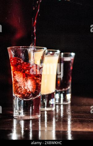 Trois verres à dose remplis de boissons colorées, l'un étant versé, placés sur un fond sombre avec des reflets sur la surface Banque D'Images