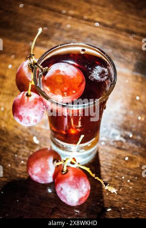 Verre à grenaille rempli de liquide rouge foncé, garni de raisins frais, posé contre une surface en bois avec des reflets légers Banque D'Images