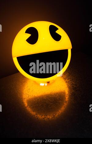 Lampe à visage smiley jaune illuminée projetant une lueur joyeuse et une ombre sur une surface texturée dans une pièce sombre Banque D'Images