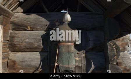 Vue arrière d'un ancien bogatyr russe en armure de chaîne et casque venant à la vieille maison en bois. Support. Concept de légendes russes et folklore Banque D'Images