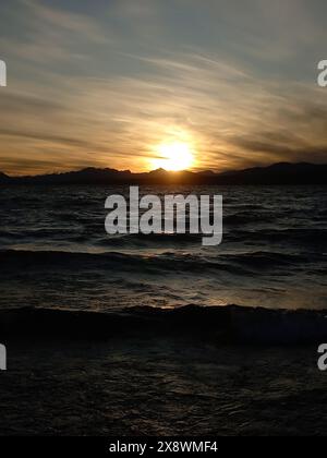 Coucher de soleil doré projetant des teintes chaudes sur un lac serein avec des vagues douces et des montagnes silhouettes au loin Banque D'Images