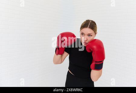 Confiant déterminé jeune femme blonde combattant, boxeur féminin exerçant avec des gants de boxe, poinçonnant en avant, isolé sur des briques blanches mur backgrou Banque D'Images