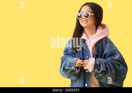 Jolie petite fille asiatique dans des vêtements adultes avec des lunettes de soleil sur fond jaune Banque D'Images