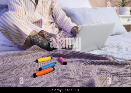 Jeune homme beau en pyjama avec différentes cigarettes électroniques jetables et ordinateur portable sur le lit à la maison Banque D'Images